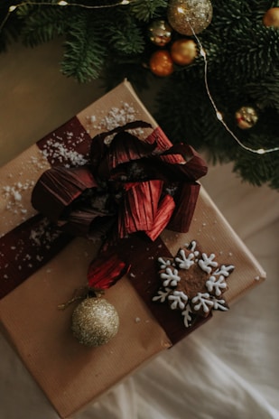 A beautifully wrapped gift with a large red bow is adorned with snowflake-shaped decorations and a gold glittery ornament. The gift is set against a backdrop of lush green fir branches and subtle string lights.