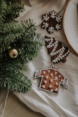 A set of holiday-themed cookies arranged on a white cloth. The cookies are shaped like a snowflake, a Christmas tree, and a sweater, all decorated with white icing. Part of an evergreen branch with a golden ornament is visible in the foreground.