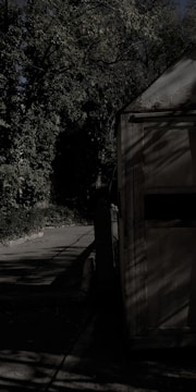 A dimly lit scene featuring a wooden structure, possibly a shed, with shadows cast across its surface. Surrounding the structure are thick trees and foliage, creating a dense and somewhat eerie atmosphere. The path nearby appears worn and slightly overgrown.
