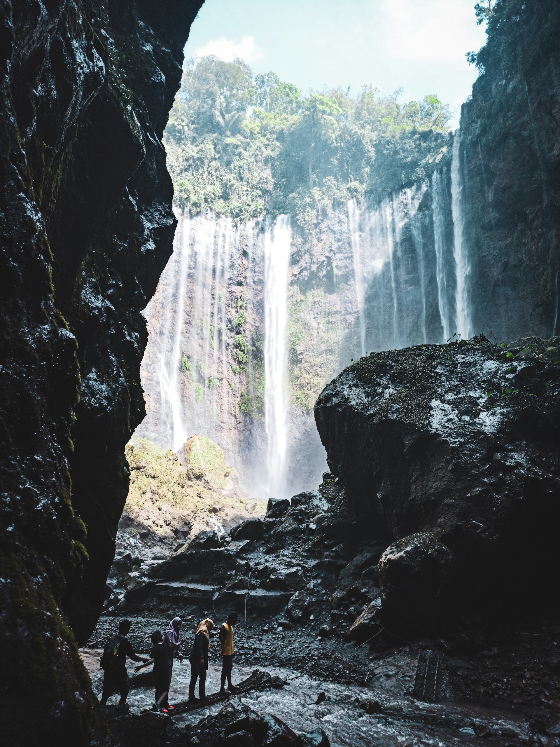 Adventurous travelers standing joyfully under a powerful waterfall surrounded by vibrant jungle.