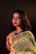 A woman with long dark hair is dressed in a patterned sari featuring floral designs. She wears heavy makeup with bold lipstick and has a bindi on her forehead. Her jewelry includes large statement pieces around her neck and a bracelet on her wrist. The background is dark, highlighting her features and outfit.