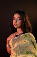 A woman with long dark hair is dressed in a patterned sari featuring floral designs. She wears heavy makeup with bold lipstick and has a bindi on her forehead. Her jewelry includes large statement pieces around her neck and a bracelet on her wrist. The background is dark, highlighting her features and outfit.