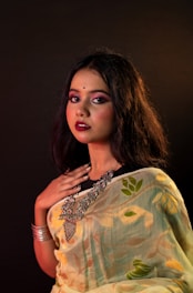 A woman with long dark hair is dressed in a patterned sari featuring floral designs. She wears heavy makeup with bold lipstick and has a bindi on her forehead. Her jewelry includes large statement pieces around her neck and a bracelet on her wrist. The background is dark, highlighting her features and outfit.