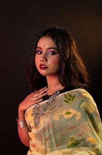 A woman with long dark hair is dressed in a patterned sari featuring floral designs. She wears heavy makeup with bold lipstick and has a bindi on her forehead. Her jewelry includes large statement pieces around her neck and a bracelet on her wrist. The background is dark, highlighting her features and outfit.