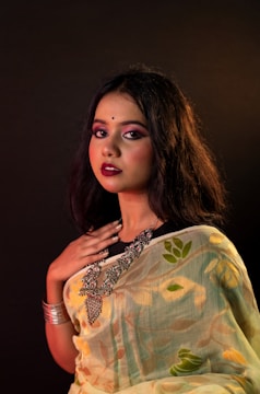 A woman with long dark hair is dressed in a patterned sari featuring floral designs. She wears heavy makeup with bold lipstick and has a bindi on her forehead. Her jewelry includes large statement pieces around her neck and a bracelet on her wrist. The background is dark, highlighting her features and outfit.