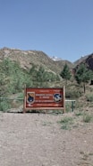 A wooden sign stands on a gravel path surrounded by grassy areas and small trees. The sign indicates a natural monument area called El Morado, welcoming visitors. Behind the sign, hills and a mountain with some snow cover are visible under a clear blue sky.