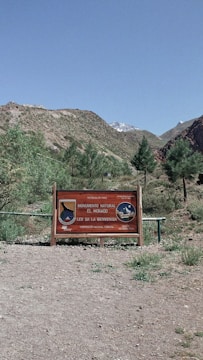 A wooden sign stands on a gravel path surrounded by grassy areas and small trees. The sign indicates a natural monument area called El Morado, welcoming visitors. Behind the sign, hills and a mountain with some snow cover are visible under a clear blue sky.