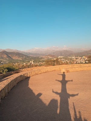 A shadow of a person with outstretched arms is cast on a dirt path under a bright blue sky. The background includes a cityscape nestled at the base of rolling mountains, visible in the distance. The warm light suggests a sunset or sunrise, creating long shadows.