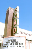 A tall, vintage-style vertical sign with large letters spelling out 'SCAD' is attached to a building. Below the sign, a marquee displays information about the Savannah Music Festival with dates ranging from March 24 to April 9, 2022. The background is a clear blue sky, suggesting a sunny day.