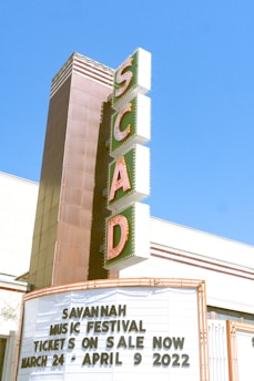 A tall, vintage-style vertical sign with large letters spelling out 'SCAD' is attached to a building. Below the sign, a marquee displays information about the Savannah Music Festival with dates ranging from March 24 to April 9, 2022. The background is a clear blue sky, suggesting a sunny day.