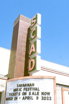 A tall, vintage-style vertical sign with large letters spelling out 'SCAD' is attached to a building. Below the sign, a marquee displays information about the Savannah Music Festival with dates ranging from March 24 to April 9, 2022. The background is a clear blue sky, suggesting a sunny day.