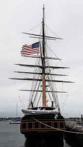 A tall ship with multiple masts docked at a harbor, displaying an American flag. The ship has intricate rigging and a classic black and brown hull. The sky is overcast, giving the scene a muted tone.