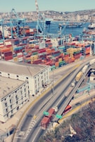 An aerial view of a busy port with numerous colorful shipping containers stacked in rows. Large blue cranes are positioned beside the containers, indicating active loading and unloading operations. In the background, the city skyline with buildings is visible across the water, where additional ships and boats are present. In the foreground, a highway with several trucks transporting containers is shown, adjacent to an industrial building.