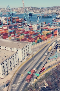 An aerial view of a busy port with numerous colorful shipping containers stacked in rows. Large blue cranes are positioned beside the containers, indicating active loading and unloading operations. In the background, the city skyline with buildings is visible across the water, where additional ships and boats are present. In the foreground, a highway with several trucks transporting containers is shown, adjacent to an industrial building.