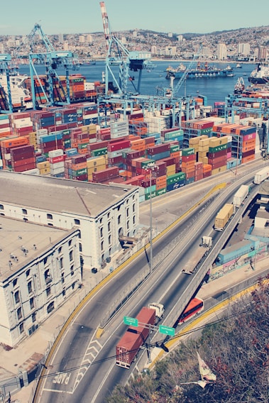 An aerial view of a busy port with numerous colorful shipping containers stacked in rows. Large blue cranes are positioned beside the containers, indicating active loading and unloading operations. In the background, the city skyline with buildings is visible across the water, where additional ships and boats are present. In the foreground, a highway with several trucks transporting containers is shown, adjacent to an industrial building.