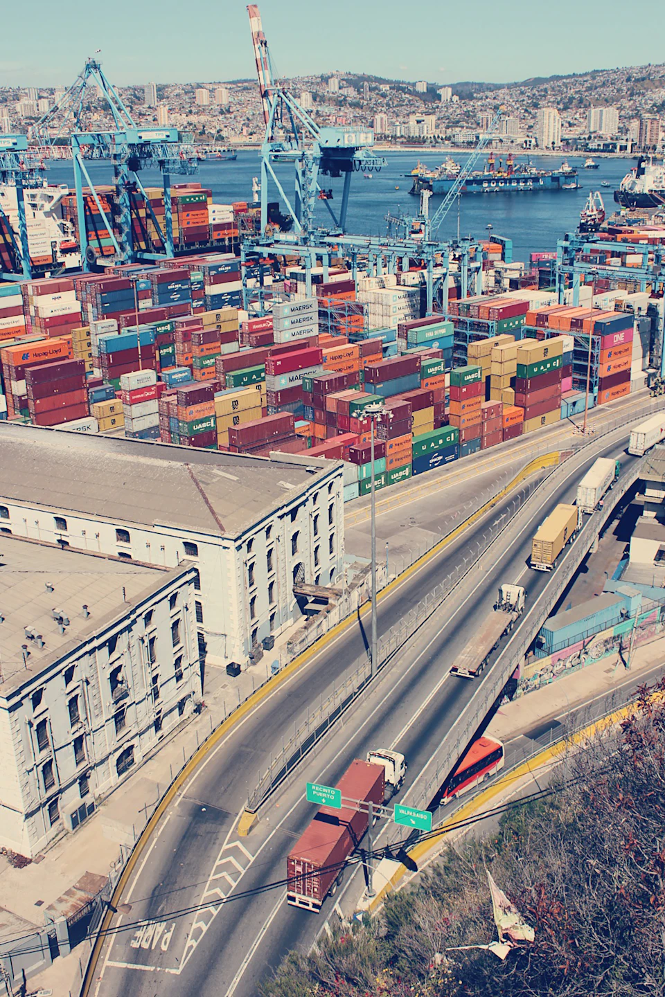 The image presents an aerial view of a bustling port city, teeming with activity and filled with cranes towering over the scene. The cityscape is dominated by numerous shipping containers stacked high on docks and cranes, indicating a thriving trade hub. A highway runs through the heart of the city, serving as a vital artery for transportation and commerce. Cars can be seen driving along this road, adding to the dynamic nature of the urban landscape. In the foreground, there are buildings that house various businesses, contributing to the overall vibrancy of the city. The sky above is a clear blue, suggesting a bright and sunny day.