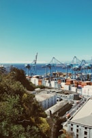 A busy port with large cranes and numerous shipping containers stacked in various colors. The scene overlooks a coastal industrial area surrounded by lush greenery and a clear view of the sea in the background. Buildings with industrial architecture and a road with traffic passing by are visible.