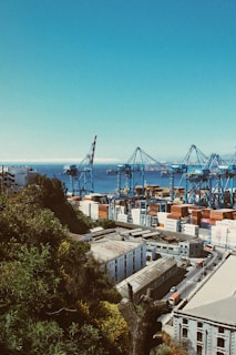 A busy port with large cranes and numerous shipping containers stacked in various colors. The scene overlooks a coastal industrial area surrounded by lush greenery and a clear view of the sea in the background. Buildings with industrial architecture and a road with traffic passing by are visible.
