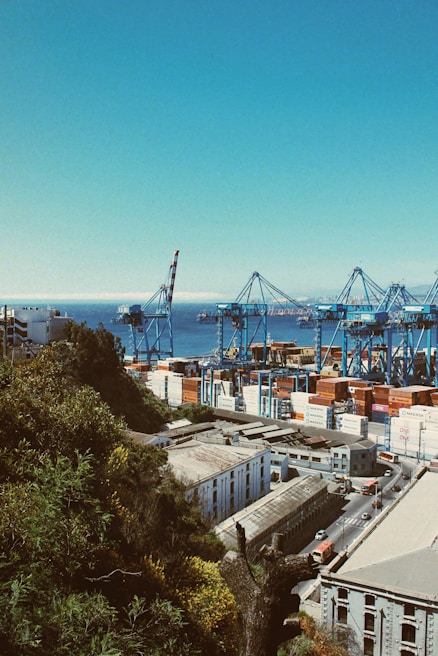 A busy port with large cranes and numerous shipping containers stacked in various colors. The scene overlooks a coastal industrial area surrounded by lush greenery and a clear view of the sea in the background. Buildings with industrial architecture and a road with traffic passing by are visible.