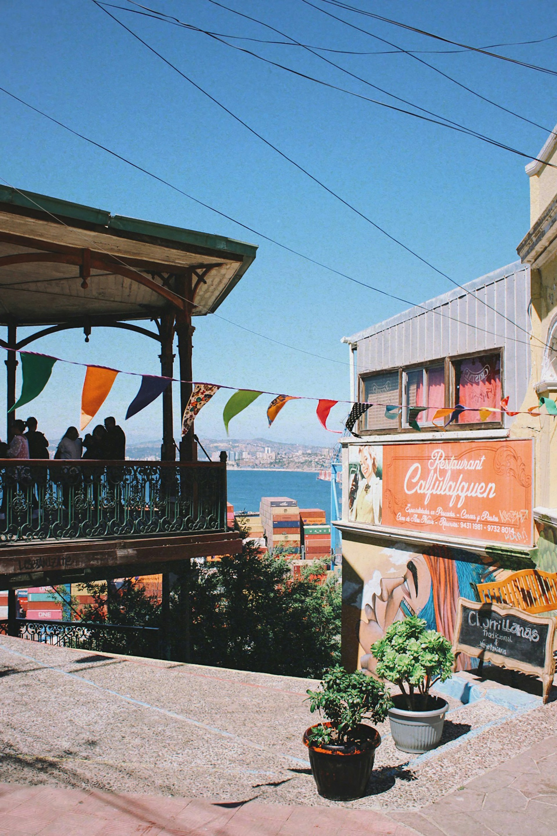 A vibrant street scene with a colorful wall mural and a sign for a restaurant named 'Café Turri'. Decorative flags are strung along a balcony overlooking the sea in the background. The area is adorned with potted plants and has a lively, artistic atmosphere.