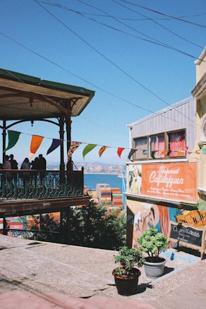 A vibrant street scene with a colorful wall mural and a sign for a restaurant named 'Café Turri'. Decorative flags are strung along a balcony overlooking the sea in the background. The area is adorned with potted plants and has a lively, artistic atmosphere.