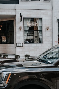 A storefront with a large window displaying the name 'Revivalist', framed by white curtains. The building exterior is made of light-colored bricks with vintage-style light fixtures on either side of the window. In front of the store, a dark-colored SUV is partially visible, reflecting its surroundings. Small bushes are arranged neatly along the edge of the sidewalk.