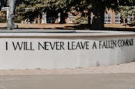 A section of a monument shows an inscription with the phrase 'I WILL NEVER LEAVE A FALLEN COMRADE' in bold capital letters. Part of a statue's leg and boot, likely of a soldier, is visible at the top left. The background features trees and a building with white-framed windows.