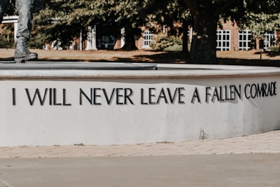 A section of a monument shows an inscription with the phrase 'I WILL NEVER LEAVE A FALLEN COMRADE' in bold capital letters. Part of a statue's leg and boot, likely of a soldier, is visible at the top left. The background features trees and a building with white-framed windows.