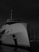 A large yacht is docked at a marina with several other masts visible in the background. The yacht's bow is prominent, creating a sense of grandeur and luxury. The image is in black and white, adding a dramatic and timeless feel.
