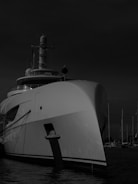 A large yacht is docked at a marina with several other masts visible in the background. The yacht's bow is prominent, creating a sense of grandeur and luxury. The image is in black and white, adding a dramatic and timeless feel.