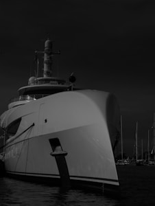 A large yacht is docked at a marina with several other masts visible in the background. The yacht's bow is prominent, creating a sense of grandeur and luxury. The image is in black and white, adding a dramatic and timeless feel.