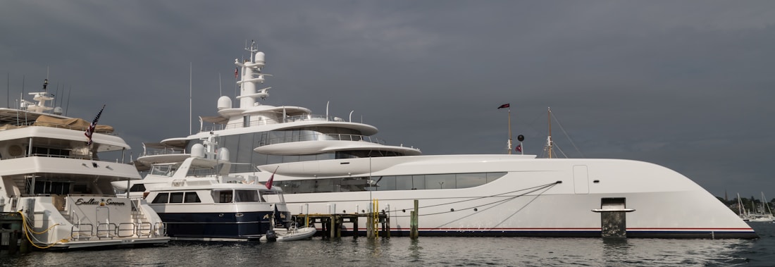 Several luxury yachts are docked at a marina under a cloudy sky. The water is calm, and the docks are lined with large, modern vessels, each with multiple decks and visible equipment such as antennas and lifeboats.