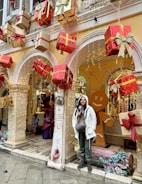 A festive display with large, wrapped gift boxes in red and gold suspended in front of an arched building entrance. A person in a white jacket stands next to a decorative column that has a message written on it. Behind them is a large gingerbread man figure and other holiday decorations.
