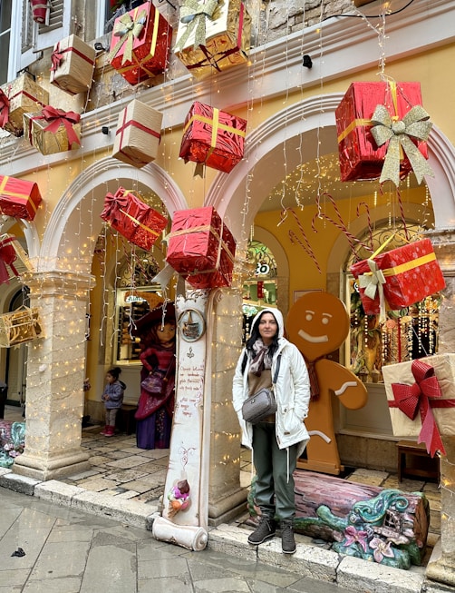 A festive display with large, wrapped gift boxes in red and gold suspended in front of an arched building entrance. A person in a white jacket stands next to a decorative column that has a message written on it. Behind them is a large gingerbread man figure and other holiday decorations.
