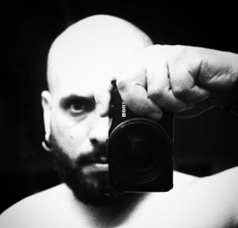 A black and white close-up photograph of a bald man with a beard holding a camera close to his face. The image has a grainy texture, adding a vintage or dramatic effect. The man's intense gaze is directed toward the camera lens, while his hand grips the device firmly.