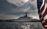 A luxurious yacht is anchored in calm waters with the sky covered in scattered clouds. The large vessel features multiple decks and modern design elements. The foreground prominently features a portion of the American flag, fluttering in the breeze.
