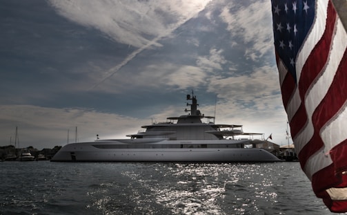A luxurious yacht is anchored in calm waters with the sky covered in scattered clouds. The large vessel features multiple decks and modern design elements. The foreground prominently features a portion of the American flag, fluttering in the breeze.