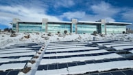 A modern building with large windows is situated behind an array of solar panels partially covered in snow. The surrounding area also has patches of snow and some rocky terrain. The sky is partly cloudy with a mix of white and blue hues.