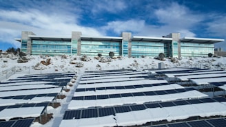 A modern building with large windows is situated behind an array of solar panels partially covered in snow. The surrounding area also has patches of snow and some rocky terrain. The sky is partly cloudy with a mix of white and blue hues.