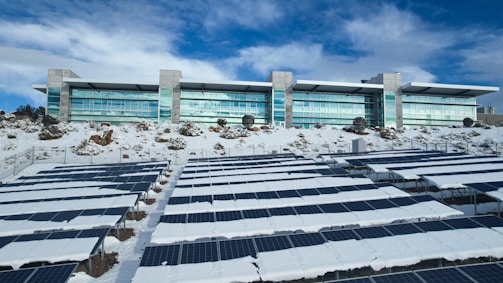 A modern building with large windows is situated behind an array of solar panels partially covered in snow. The surrounding area also has patches of snow and some rocky terrain. The sky is partly cloudy with a mix of white and blue hues.