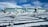 A modern building with large windows is situated behind an array of solar panels partially covered in snow. The surrounding area also has patches of snow and some rocky terrain. The sky is partly cloudy with a mix of white and blue hues.