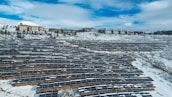 A large array of solar panels is installed across a snow-covered landscape, with modern buildings visible in the background. The sky is partly cloudy, adding a serene atmosphere.