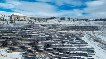 A large array of solar panels is installed across a snow-covered landscape, with modern buildings visible in the background. The sky is partly cloudy, adding a serene atmosphere.