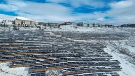A large array of solar panels is installed across a snow-covered landscape, with modern buildings visible in the background. The sky is partly cloudy, adding a serene atmosphere.