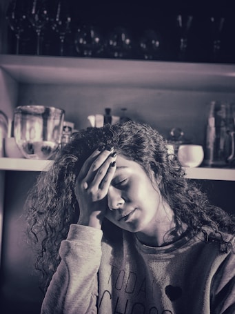 A woman with curly hair appears to be experiencing a moment of stress or fatigue, resting her forehead on her hand. She is wearing a casual sweater, and the background features shelves with glasses and kitchenware.