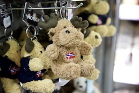 A collection of plush moose keychains hanging from a metal display rack. Each moose is small, with brown fur and large fabric antlers. They are adorned with clothing featuring a flag emblem. The background shows a slightly blurred environment, suggesting a shop setting.