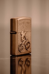 A metallic, bronze-colored lighter is intricately designed with the embossed image of a cyclist riding a bicycle. The word 'CLEVELAND' appears above the cyclist, and the lighter is positioned upright, reflecting on the glossy surface it rests upon. The background is a soft blur of warm tones.