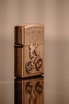 A metallic, bronze-colored lighter is intricately designed with the embossed image of a cyclist riding a bicycle. The word 'CLEVELAND' appears above the cyclist, and the lighter is positioned upright, reflecting on the glossy surface it rests upon. The background is a soft blur of warm tones.