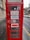 A traditional red telephone booth with signs indicating 'FOR SALE TO LET' along with contact details for inquiries. The booth is positioned on a rainy street, next to a stone building, with vehicles parked in the background.
