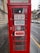 A traditional red telephone booth with signs indicating 'FOR SALE TO LET' along with contact details for inquiries. The booth is positioned on a rainy street, next to a stone building, with vehicles parked in the background.